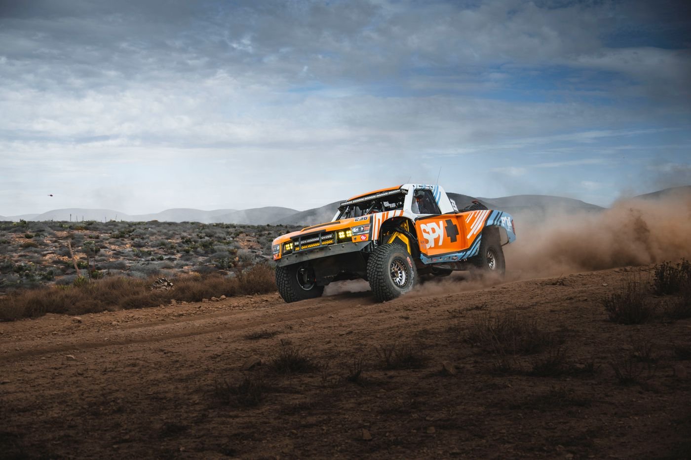 Orange and white Trophy Truck racing across Baja desert dirt, kicking up a dust trail