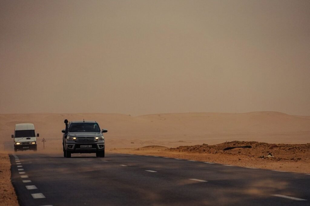 A pickup truck and a van driving along a long desert road in dusty orange light.