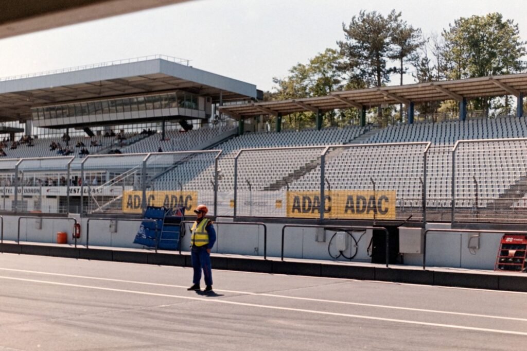 A track marshal in a high-visibility vest standing in front of empty spectator stands at a racing circuit.