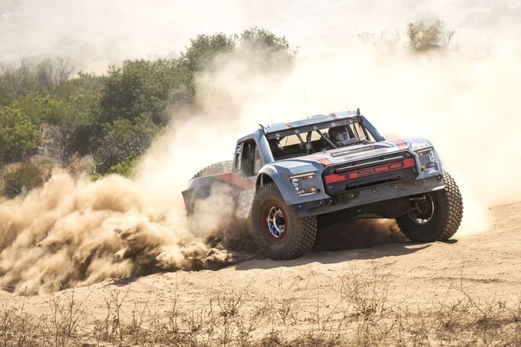 A grey and red off-road trophy truck powering through sand with dust and green scrub behind.